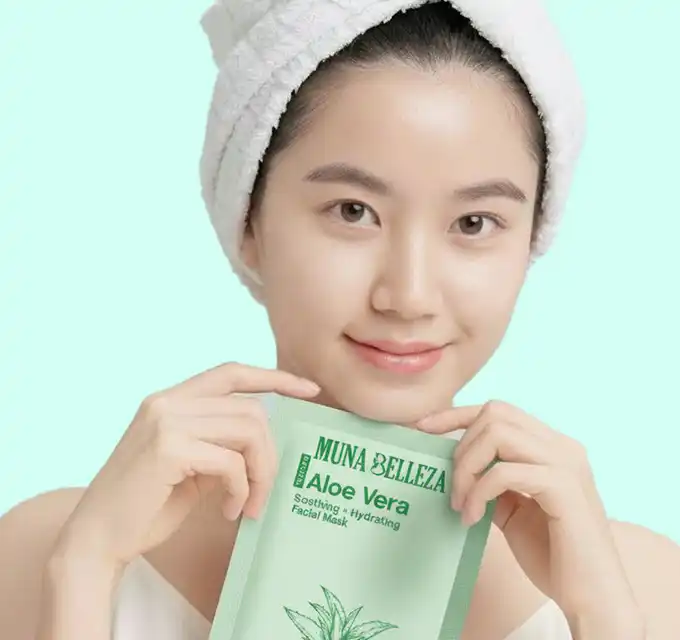 Woman wearing a towel turban presenting a green aloe vera mask packet with a soft smile and teal graphic elements behind her.